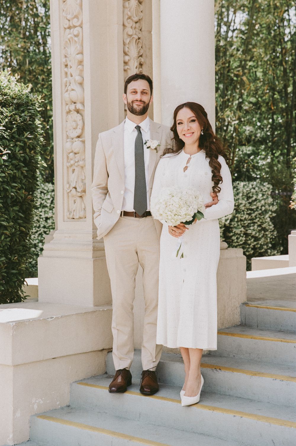A couple poses together in formal white attire against ornate white columns and greenery.