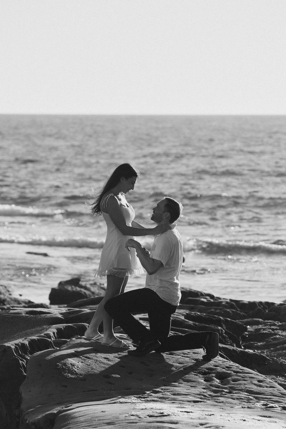 Black and white photo of a man on one knee proposing to a woman on rocky beach with ocean waves in background.