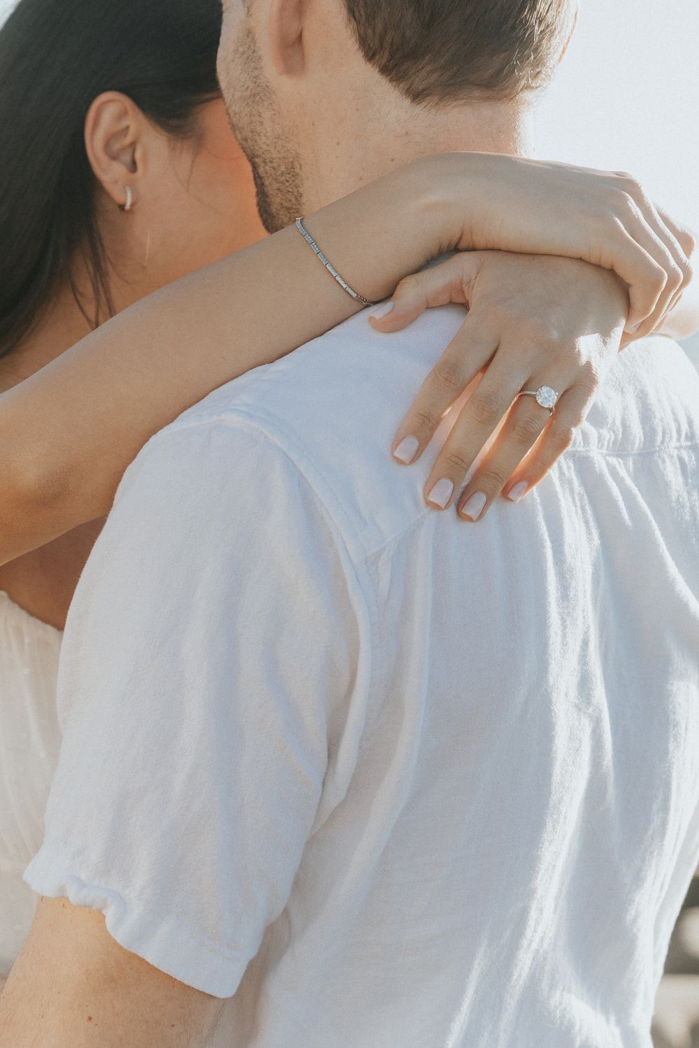 Close-up of a woman's hand with an engagement ring resting on a man's shoulder as they embrace.