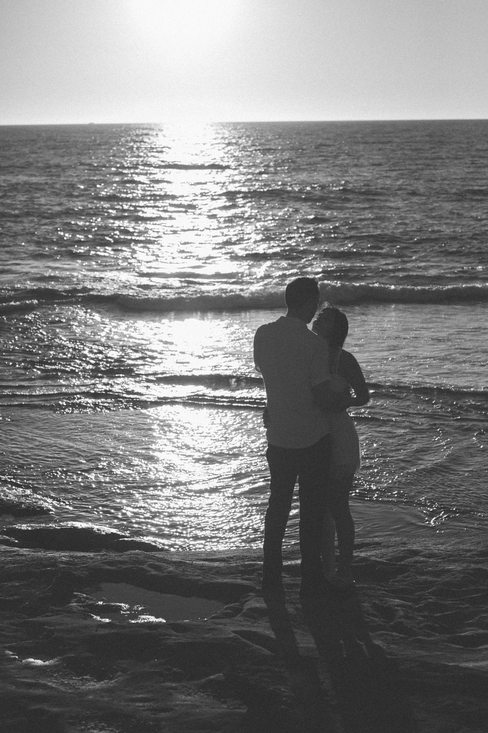 Black and white silhouette of a romantic couple embracing on a beach at sunset with ocean waves.