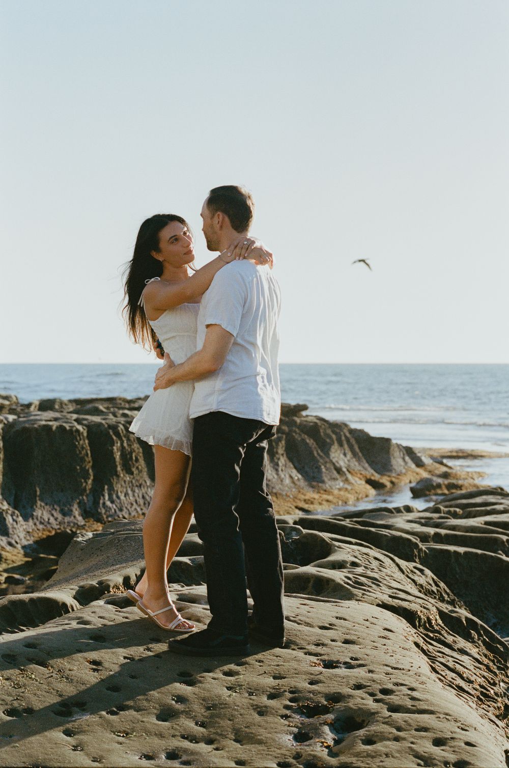 Couple embracing on rocky coastal cliffs with clear blue sky and ocean in the background.