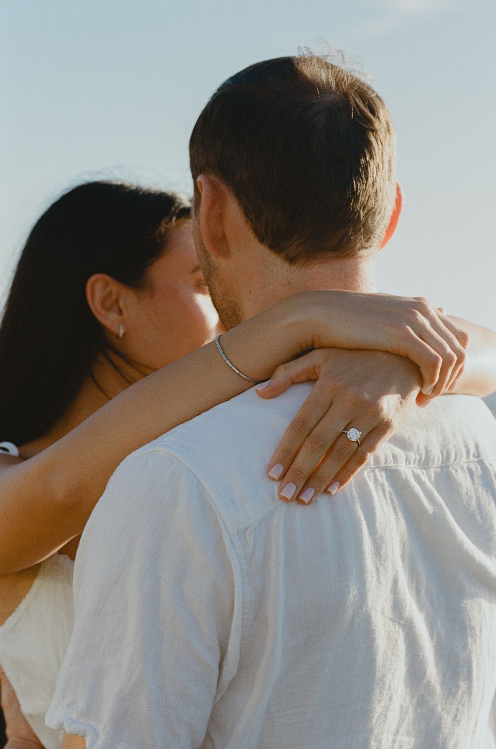 A couple embraces outdoors, the woman showing an engagement ring while hugging her partner under a clear blue sky.