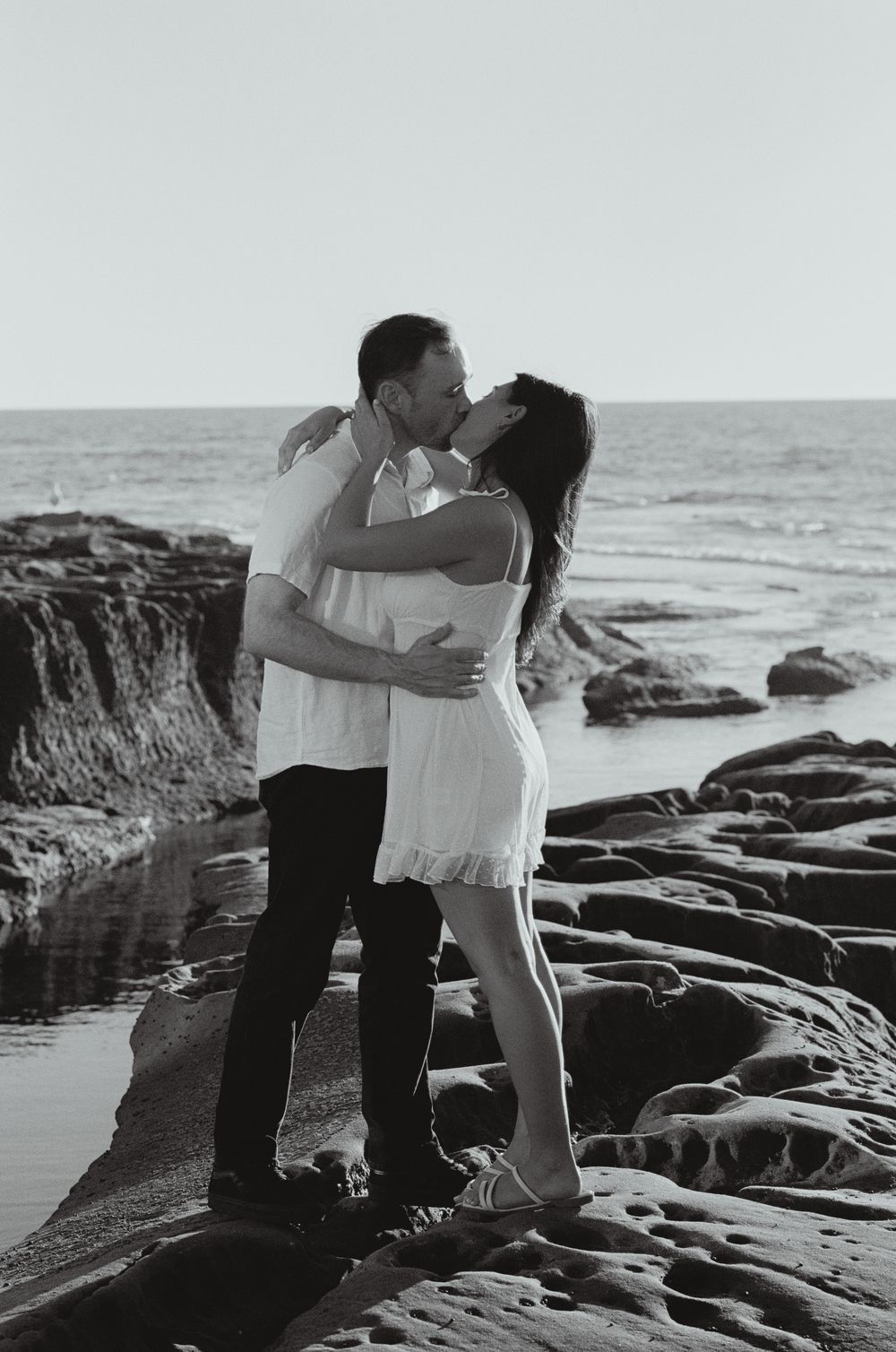 Black and white photo of a couple kissing on rocky beach shore with ocean waves in the background.