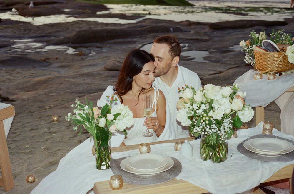 Couple sharing a romantic beach picnic at a white-draped table adorned with white floral arrangements.