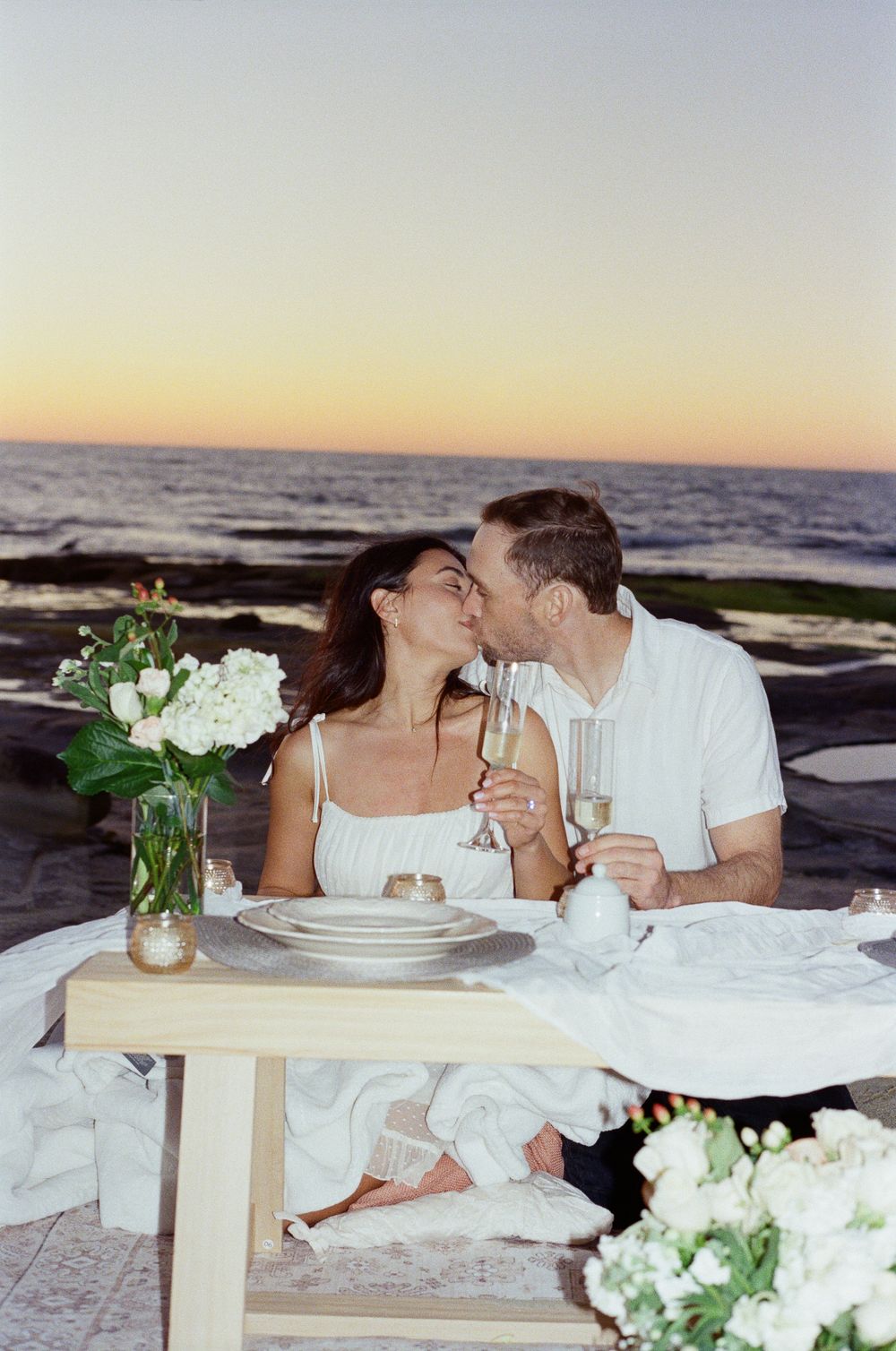 Couple sharing a romantic beachside picnic kiss at sunset with champagne and white flowers.