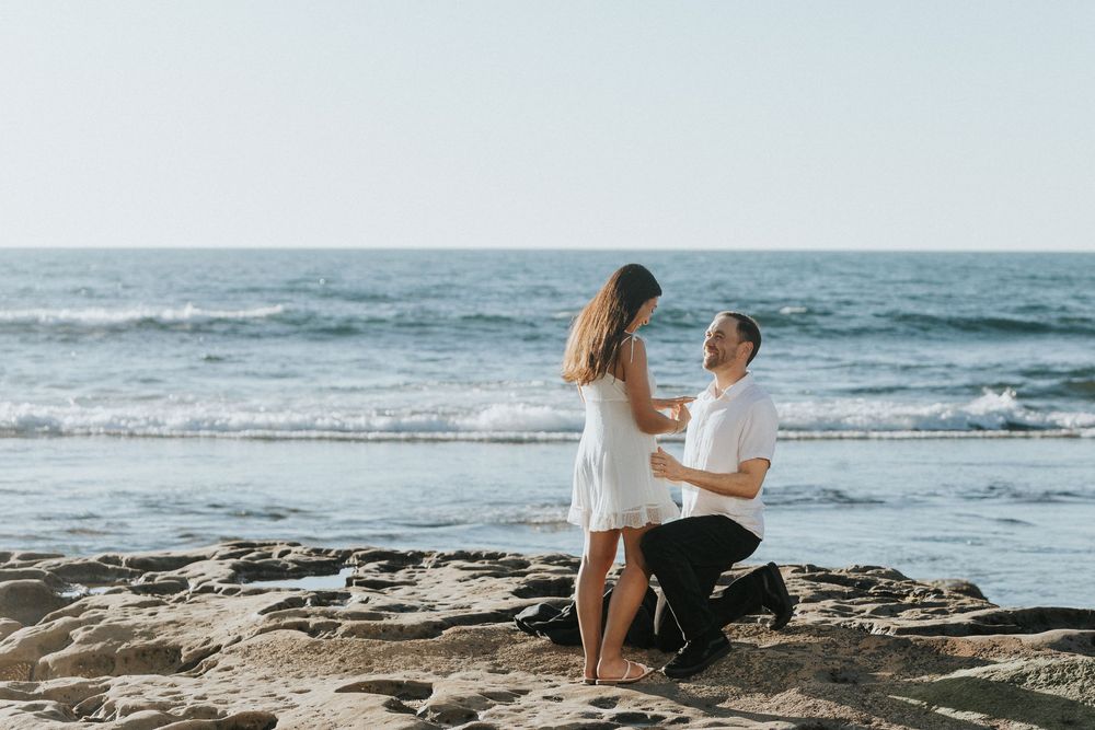Man proposing to woman on rocky beach with ocean waves in background on sunny day.