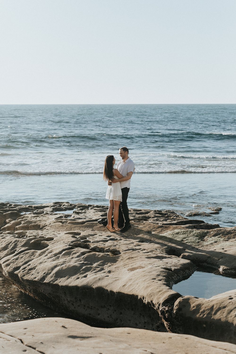 Couple embracing on rocky beach shoreline with ocean waves in background on a sunny day.