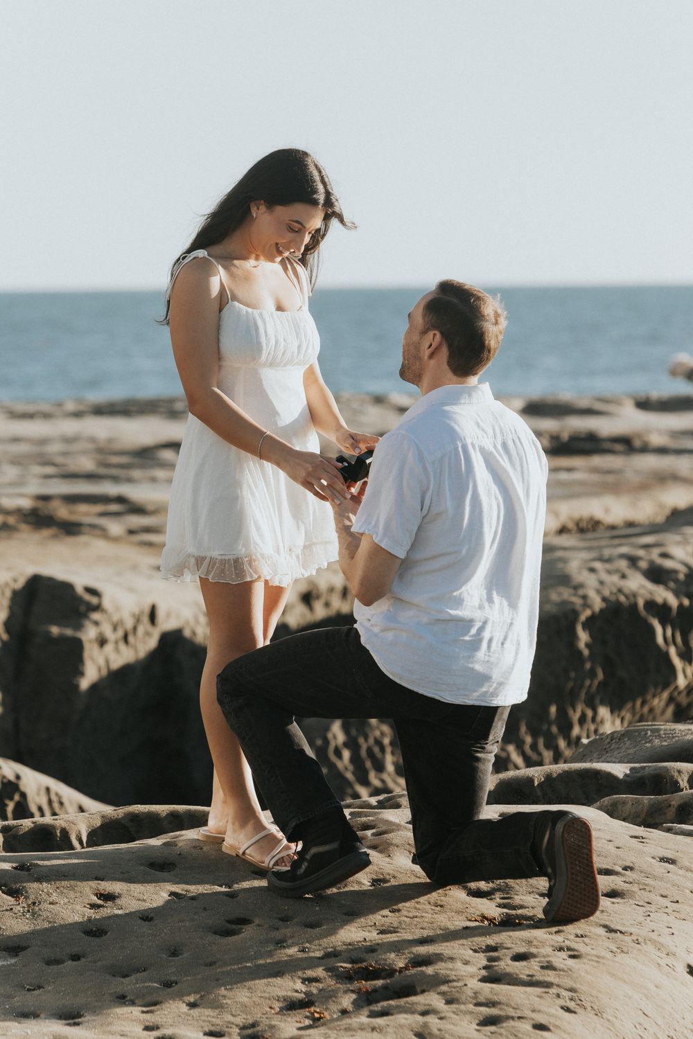 Man kneeling on rocky beach proposing to woman in white dress with ocean backdrop.