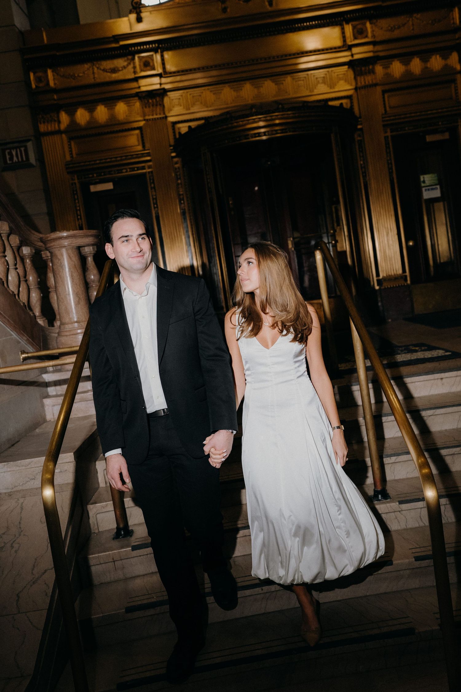 A couple in formal wedding attire walks through a dimly lit hallway with dramatic lighting and shadows.