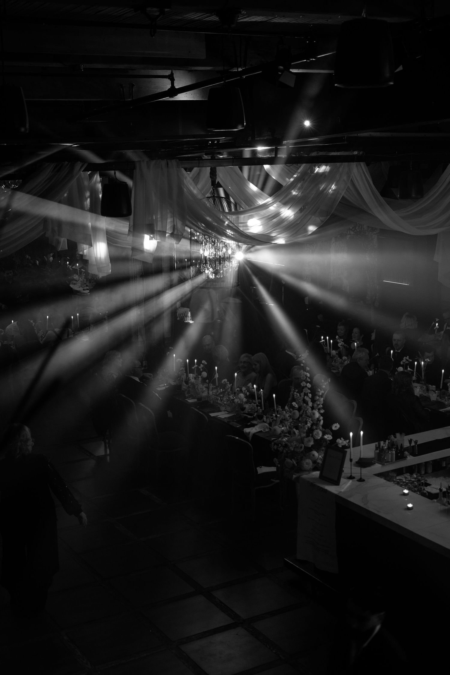 Dramatic beams of light pierce through fog and draping in a dark concert venue creating an atmospheric black and white scene.