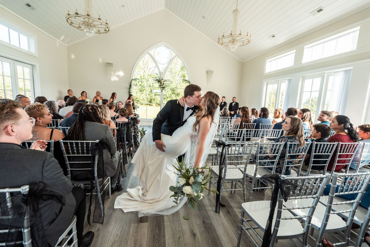 A romantic moment captured during a wedding ceremony in an elegant white chapel with gray chiavari chairs and chandeliers.