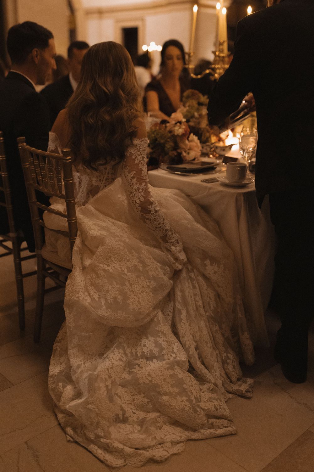 Elegant wedding reception scene showing a flowing white lace dress against dark formal attire in warm candlelight.
