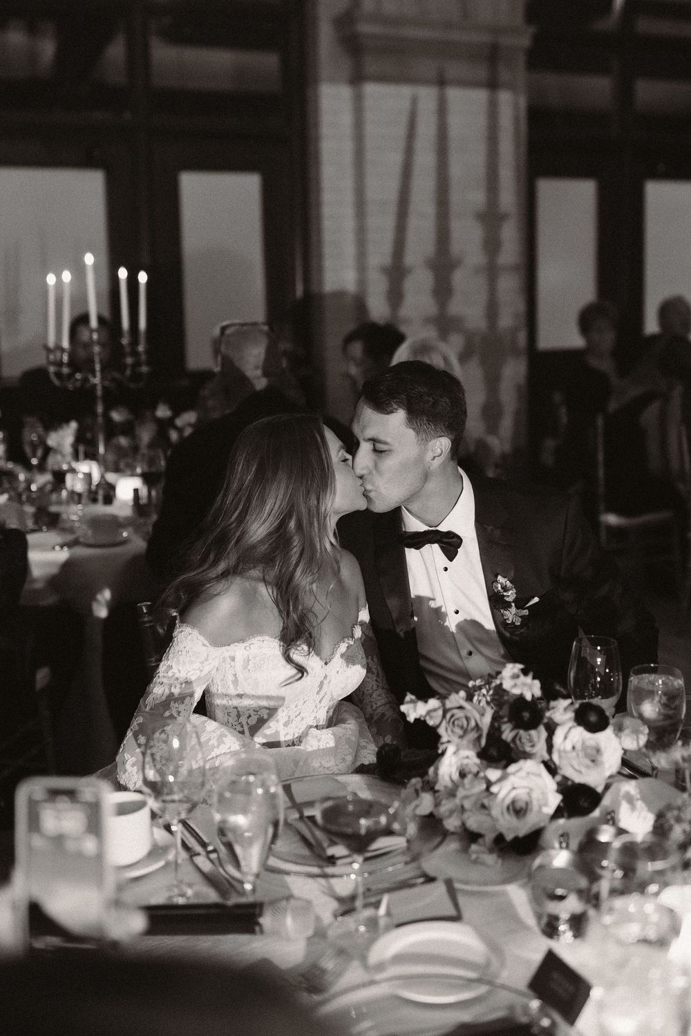 Black and white photograph of an intimate wedding reception dinner with candlelit table settings and floral centerpieces.