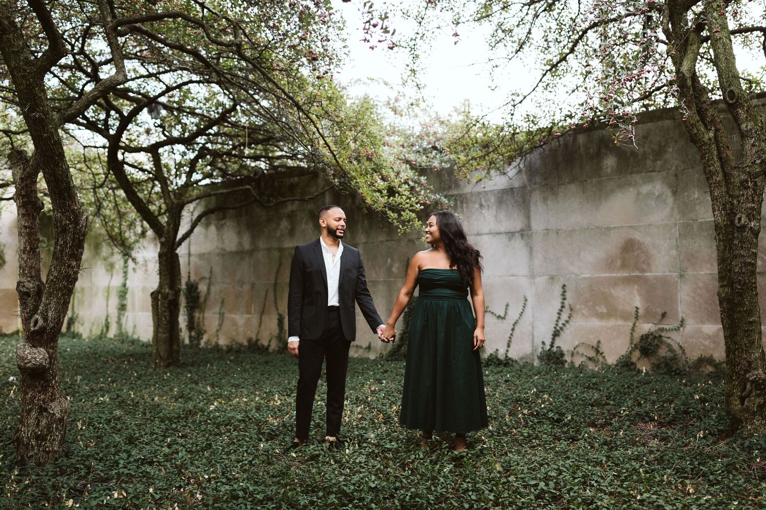 A couple poses together in a romantic garden setting surrounded by lush greenery and trees.