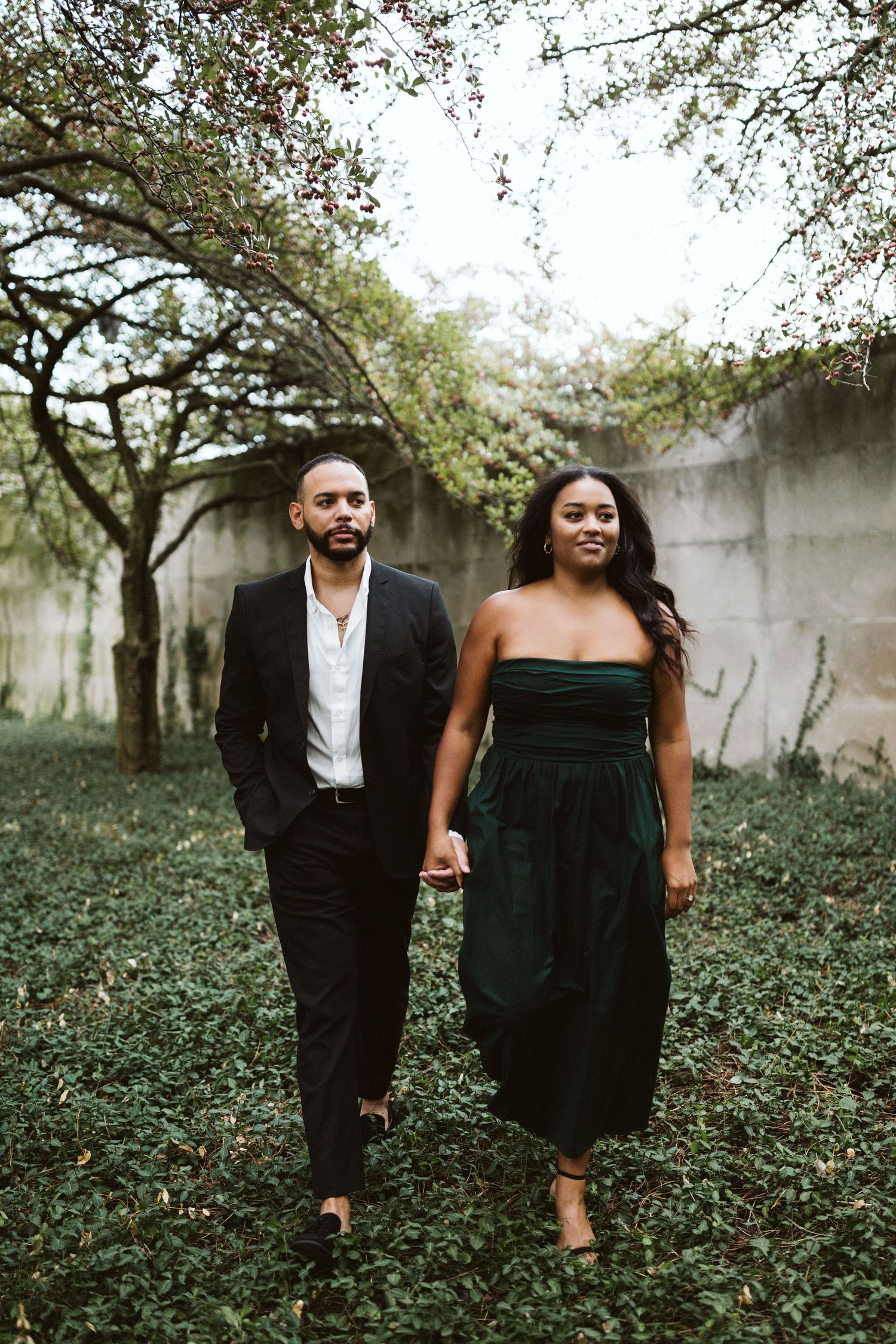 A couple in formal attire walks together through a garden setting with trees and greenery.
