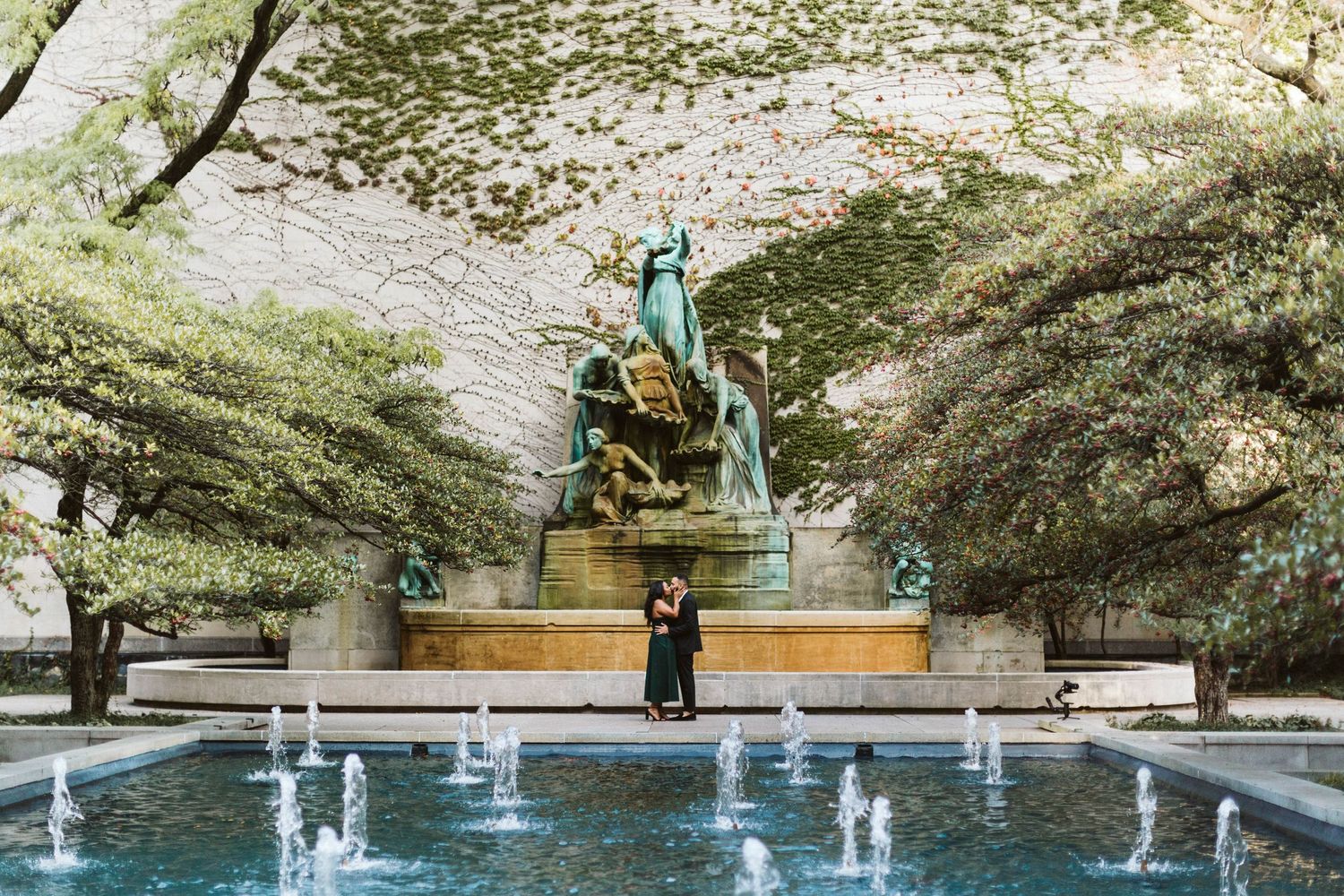 Peaceful park setting with stone benches and water fountains surrounded by lush green trees and landscaping.