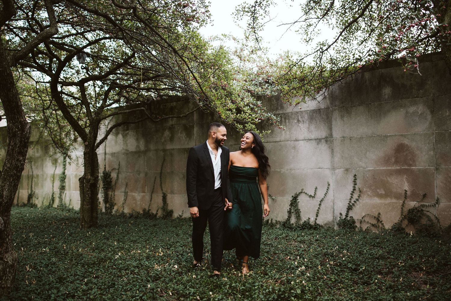 A couple in formal attire stands together against a concrete wall with overhanging tree branches in a moody setting.