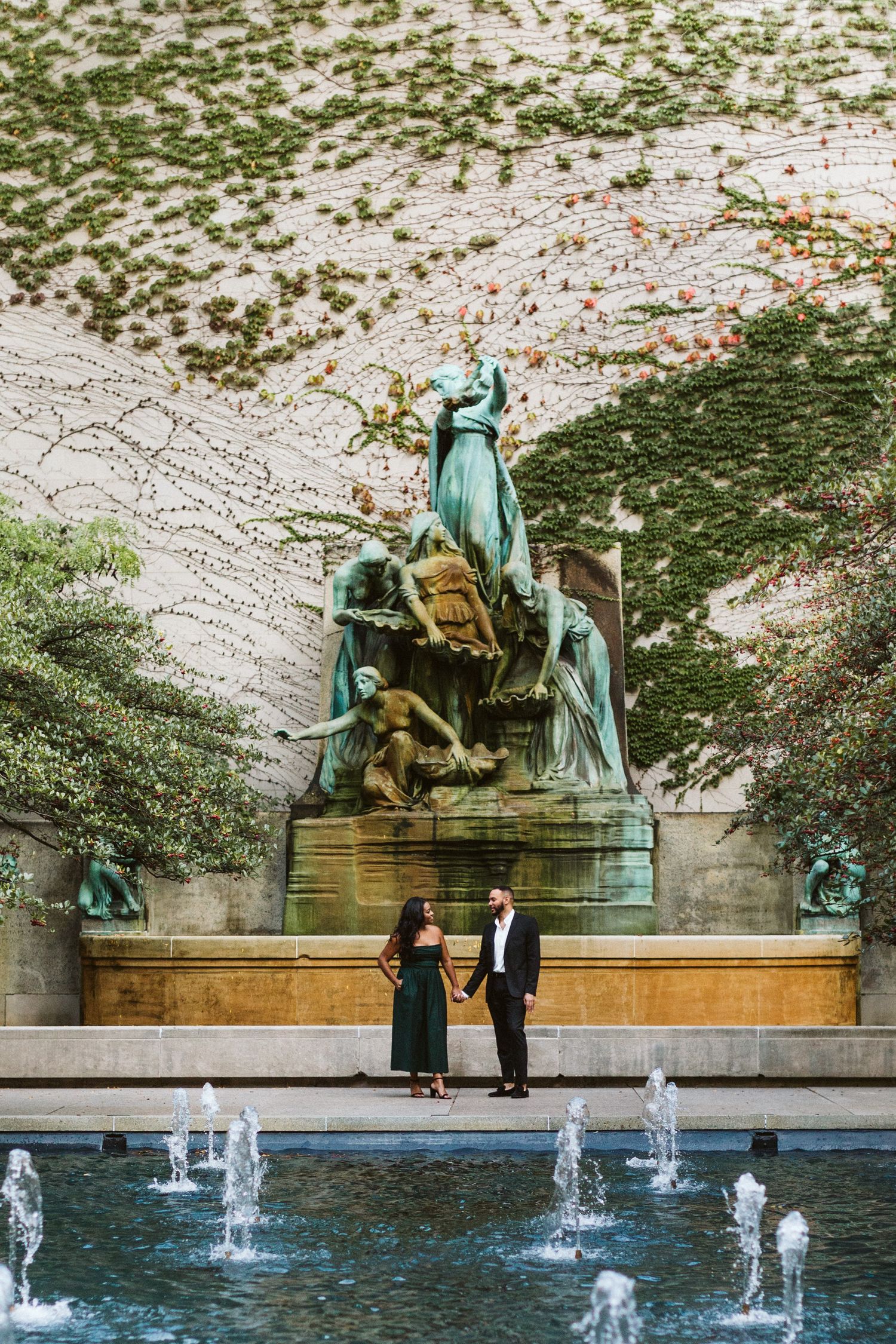 A couple stands by a grand fountain with a large bronze statue and water features in an elegant outdoor setting.