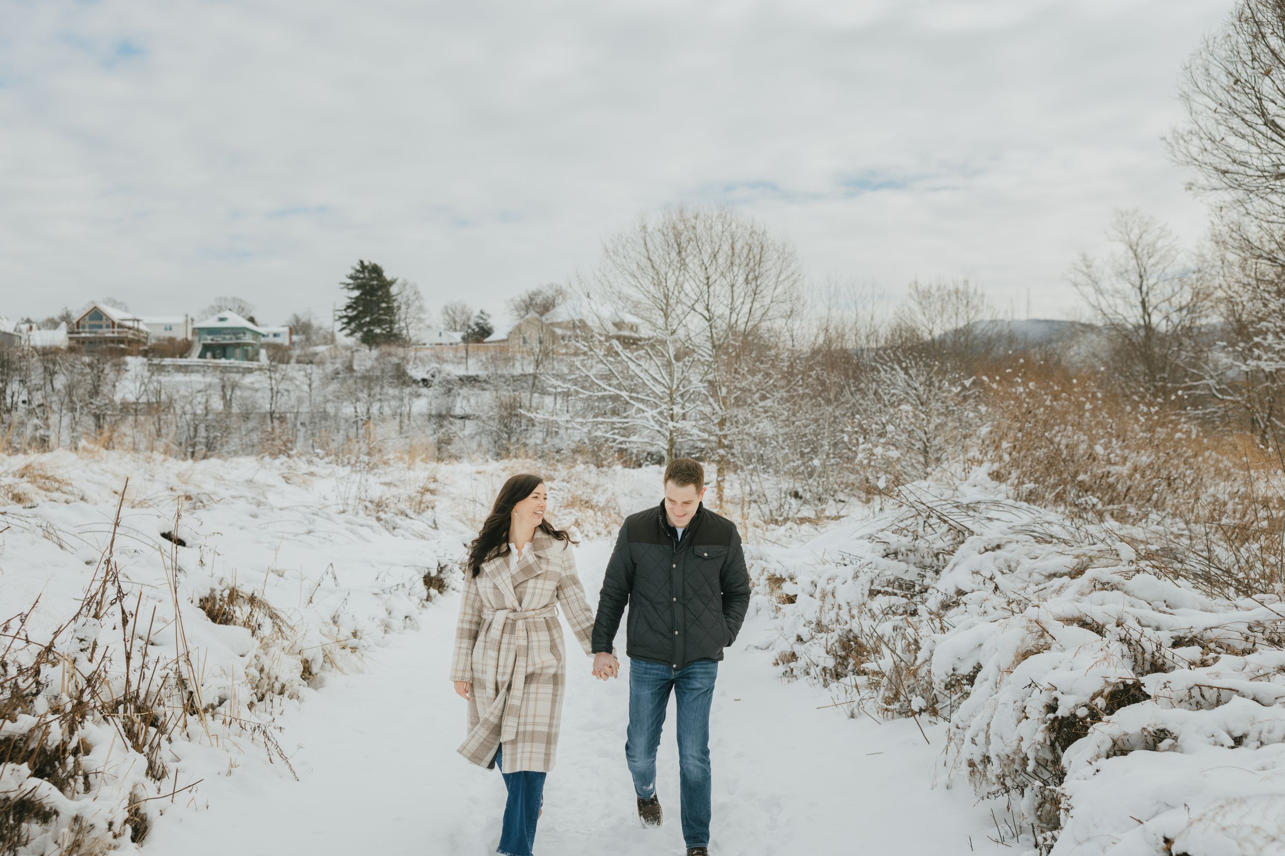 A couple walks hand in hand through a snowy winter landscape surrounded by bare trees and white ground.