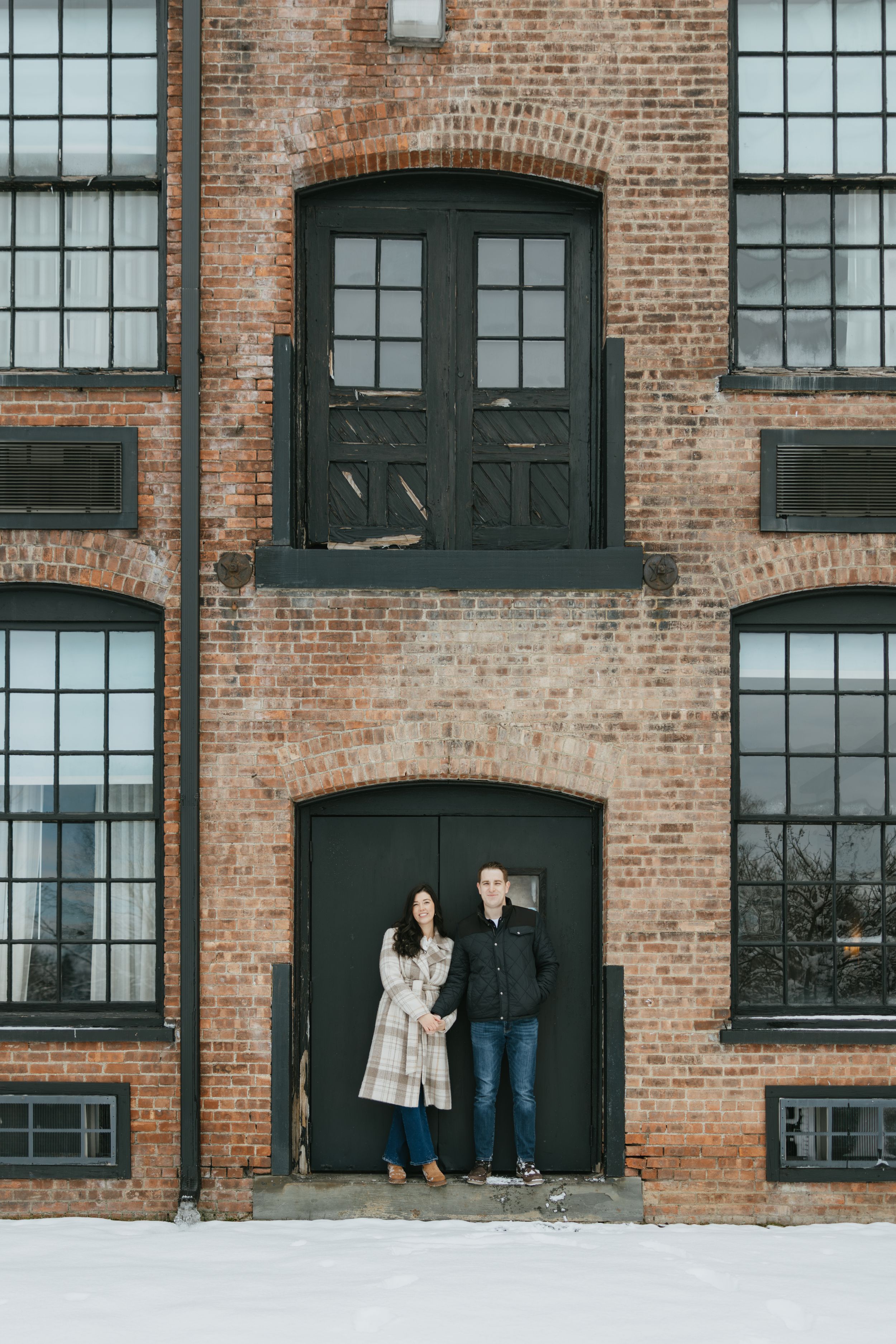 Couple poses against industrial brick building with arched doorways and windows in winter setting.