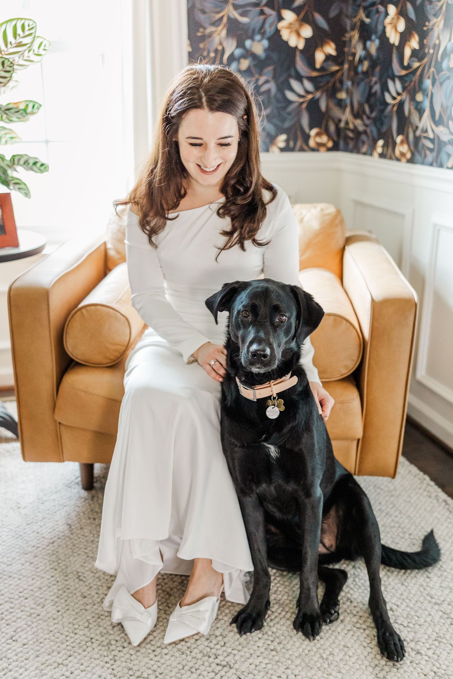 A person in a white dress sits in a tan leather chair with their black dog companion.