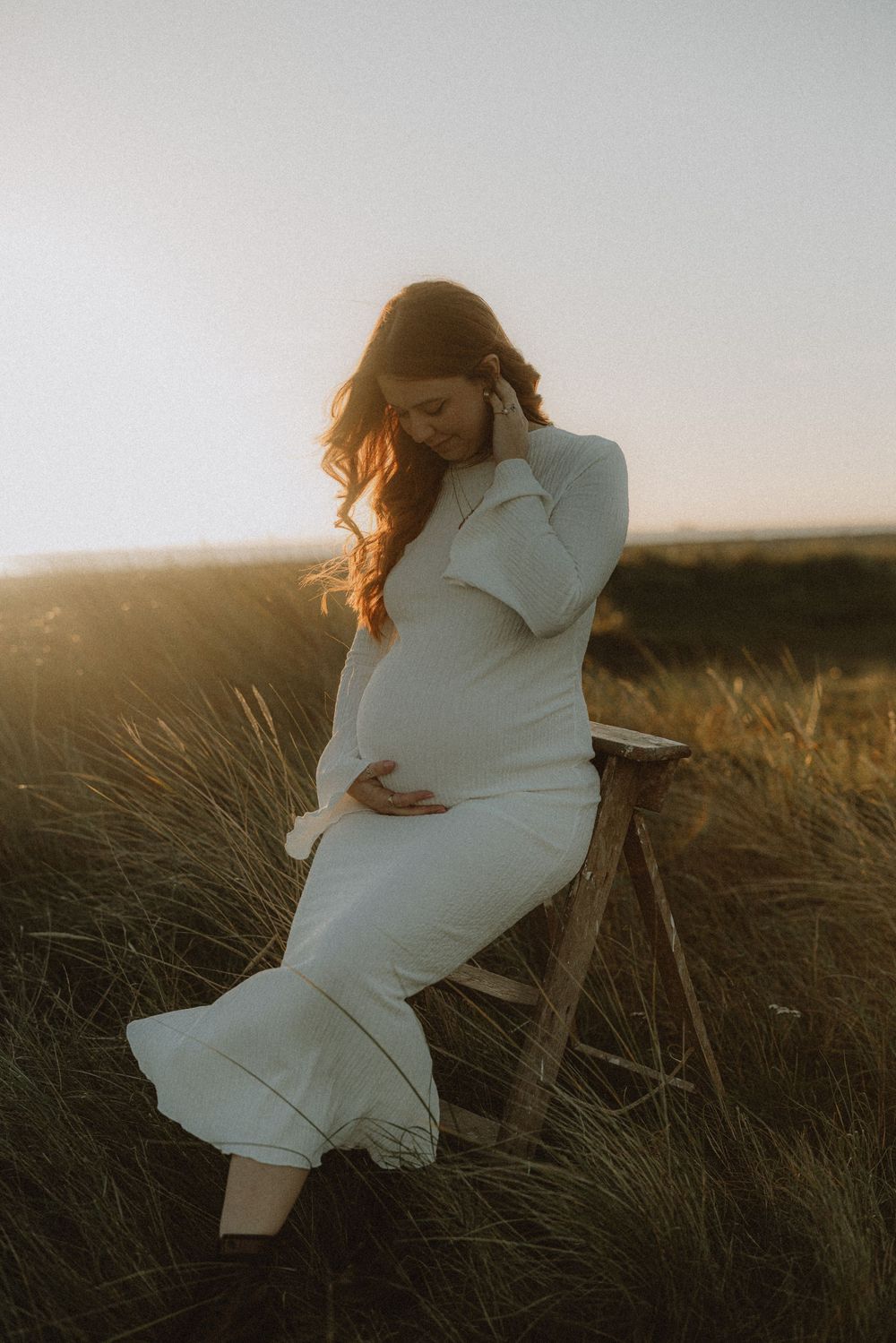 Figure in white dress poses gracefully in tall grass during golden hour sunset.