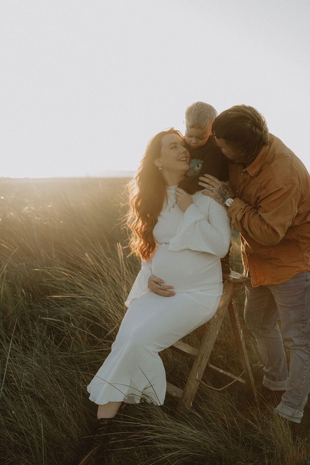 A romantic couple sits together in tall grass during golden hour sunset, creating an intimate silhouette against the sky.