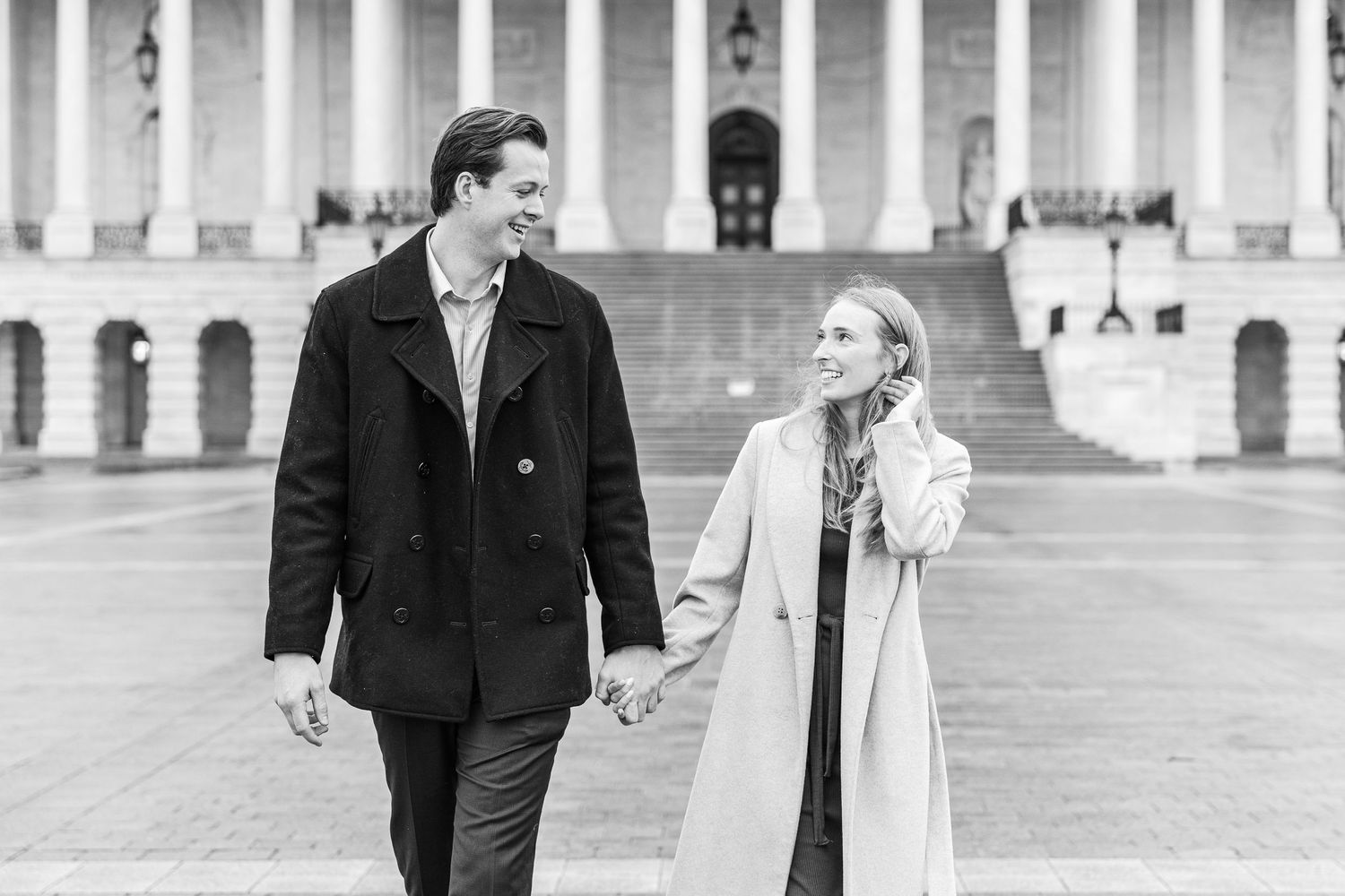 Couple in winter coats walking together in front of neoclassical government building in black and white photo.