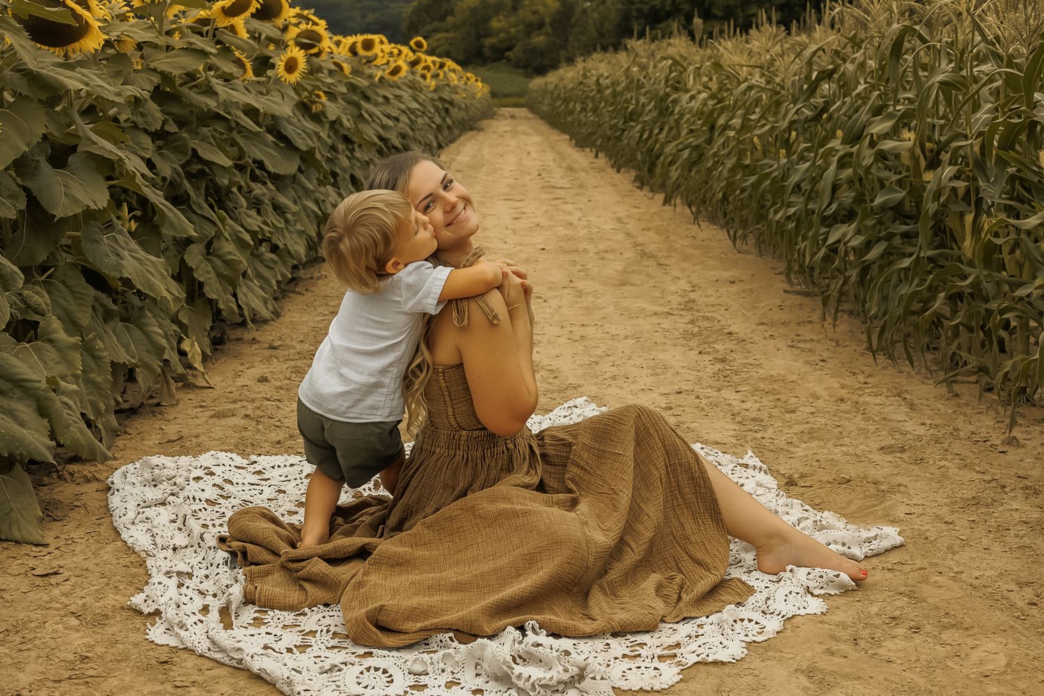 A couple in brown attire sits on a white lace blanket on a dirt path surrounded by green foliage.