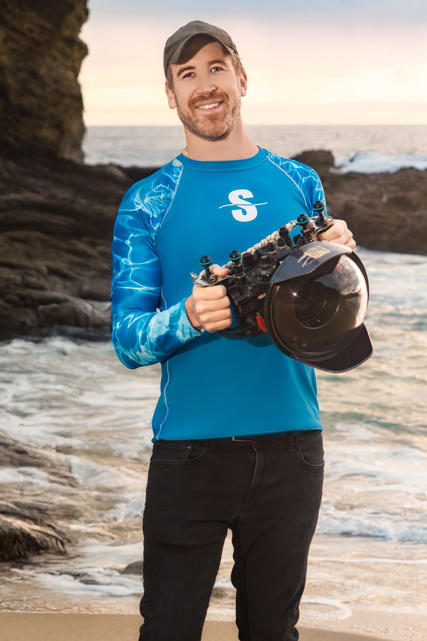 Person in bright blue sweater holding camera equipment on rocky beach at sunset.