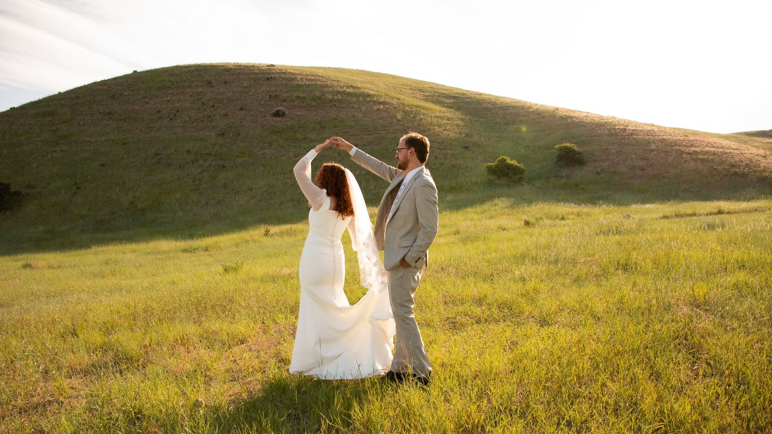 A couple dances together in a flowing white dress and suit on a sunlit grassy hillside at golden hour.