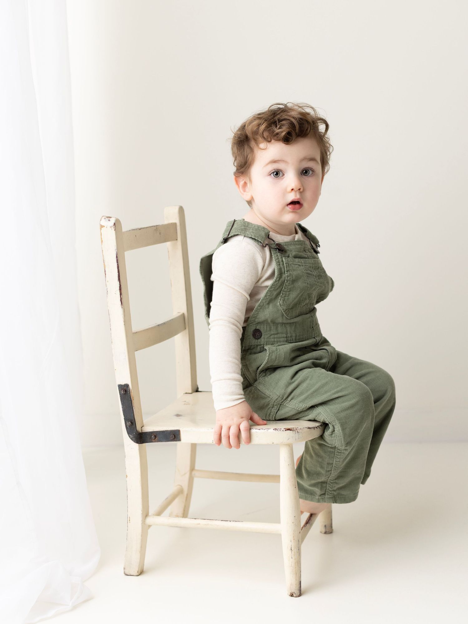 A small child in a green overalls sits and plays on a simple wooden chair against a white backdrop.