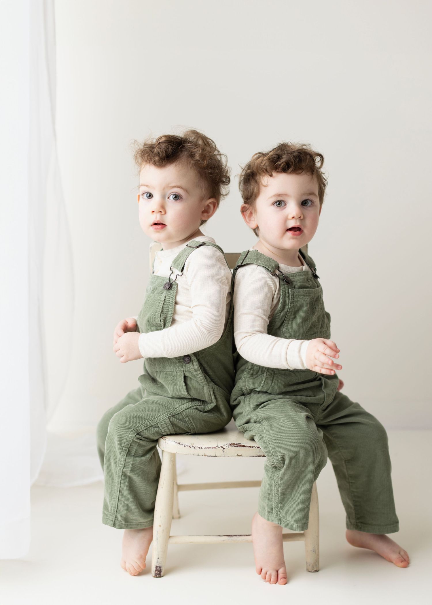 Two toddlers in matching green overalls and white shirts sit back-to-back on a small wooden chair in a bright room.