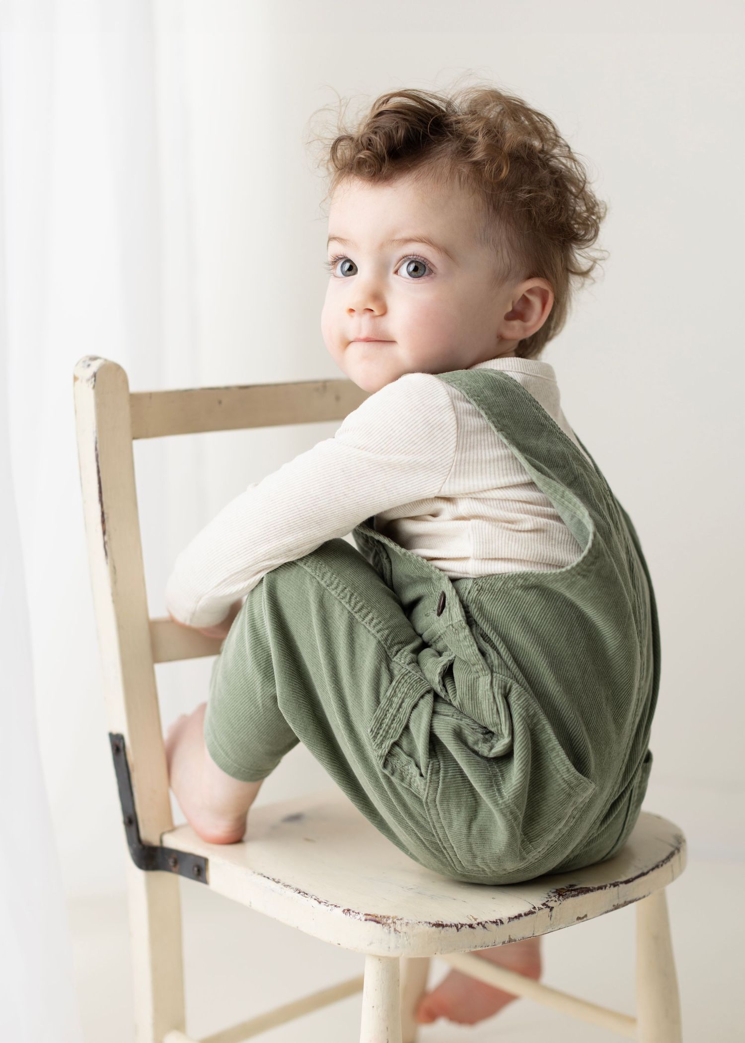 Baby in green sweater lounges playfully on white wooden chair against neutral background.