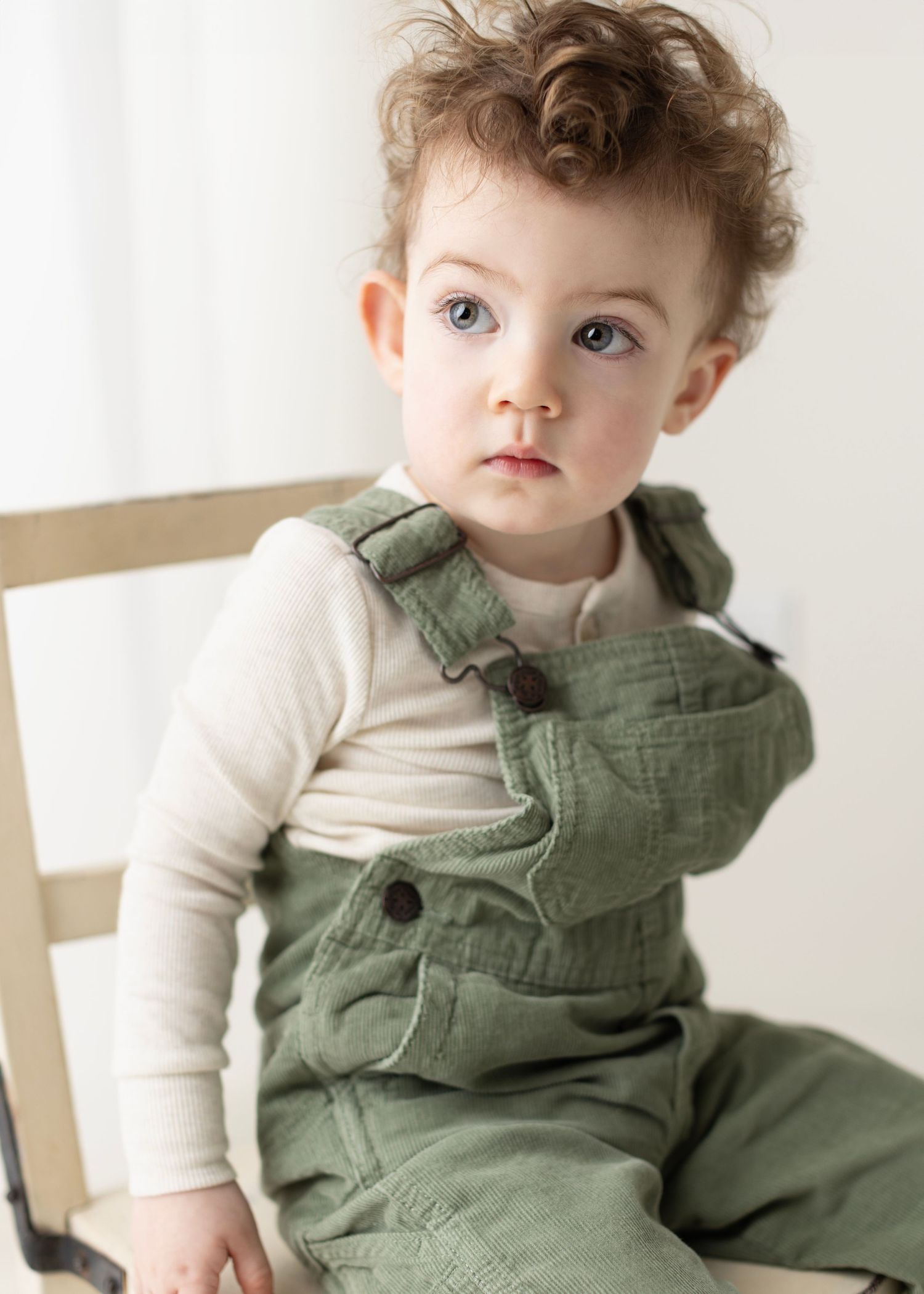Baby wearing white long sleeve shirt and olive green overalls sits on wooden chair in natural light setting.