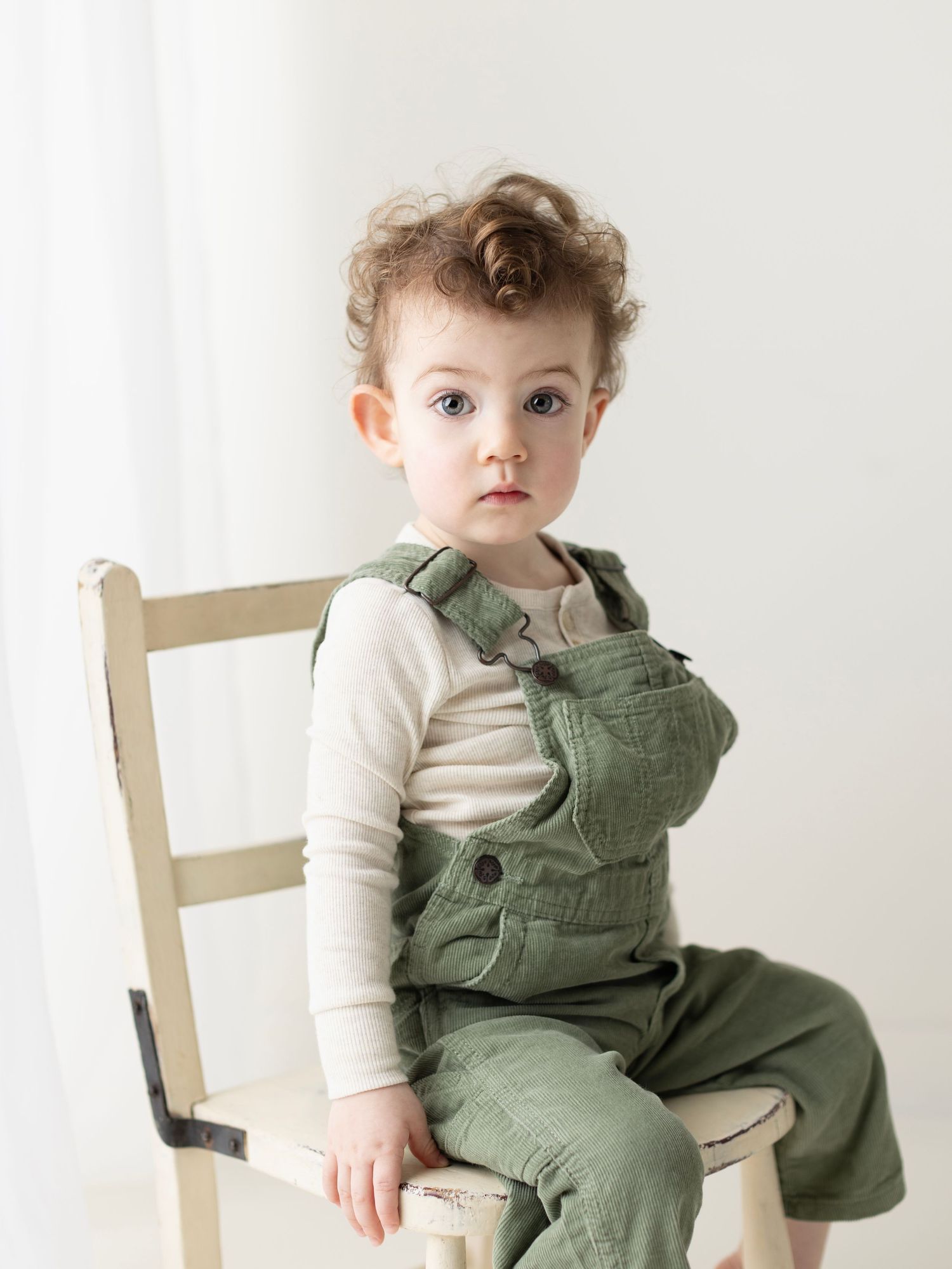 Small child wearing a cozy white long sleeve shirt and olive green overalls sits on a white wooden chair.