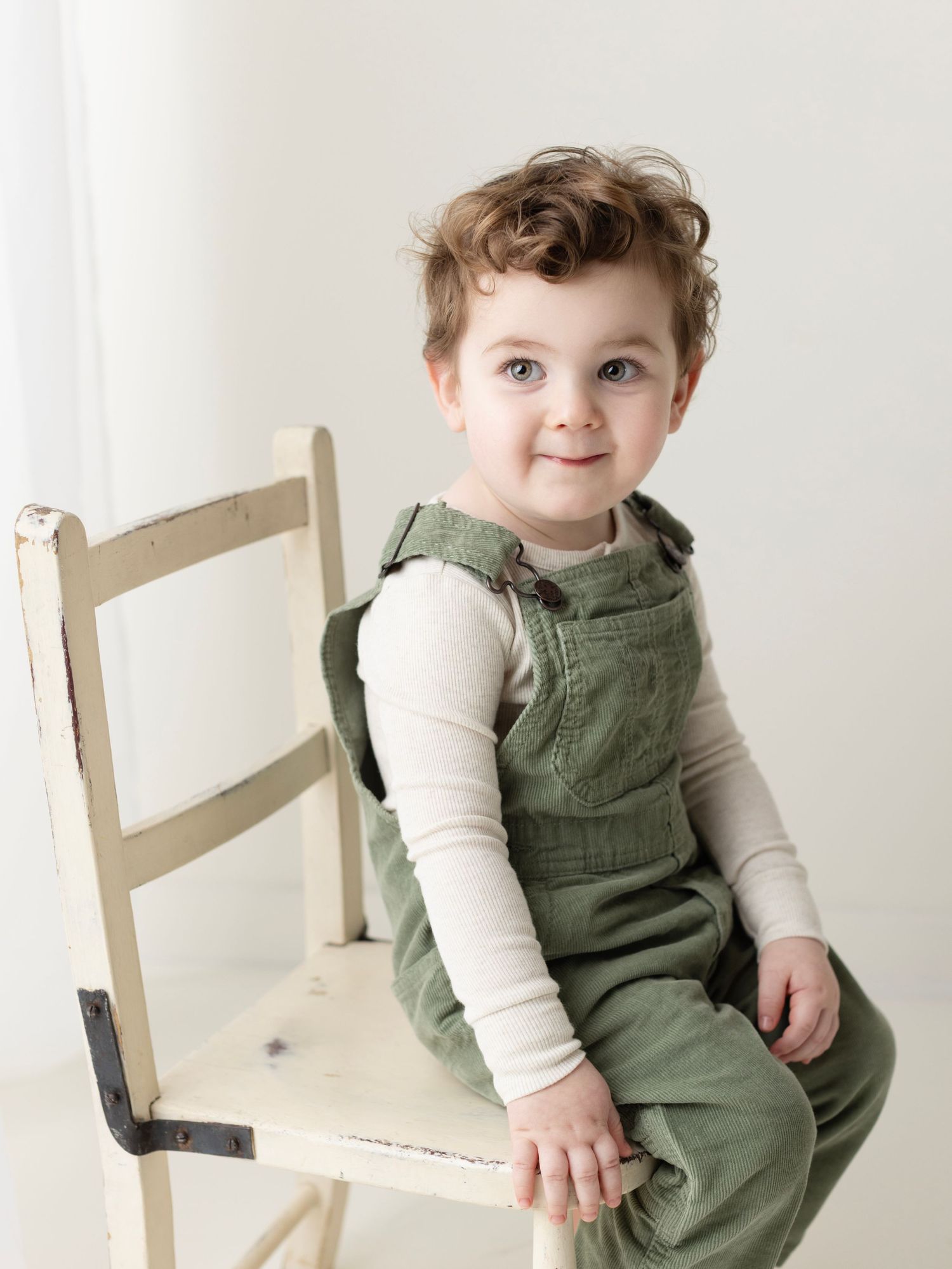 A small child wearing green overalls and a cream colored long sleeve shirt sits sideways on a white wooden chair.