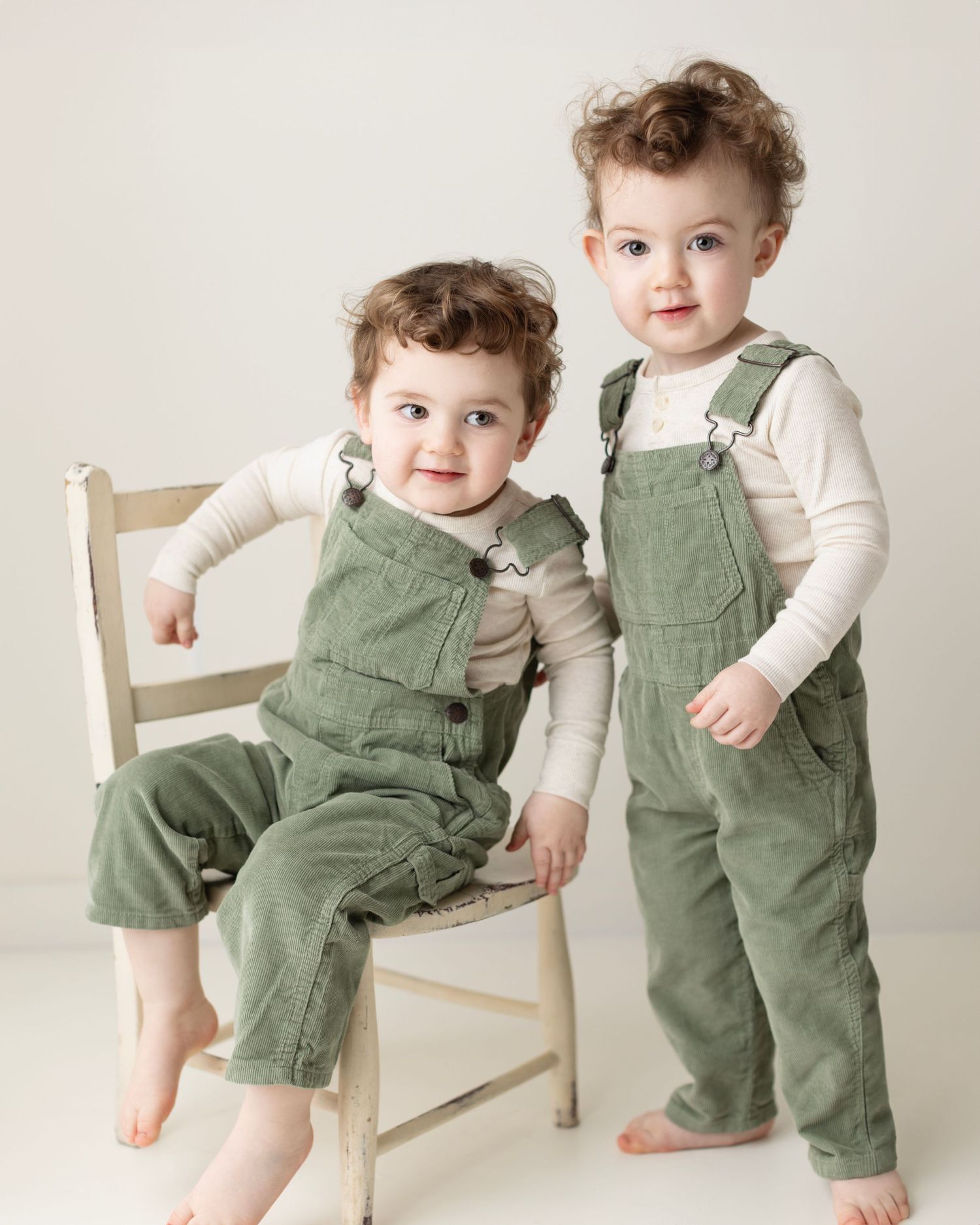 A toddler in green corduroy overalls and white shirt plays on a white wooden chair in a series of candid poses.