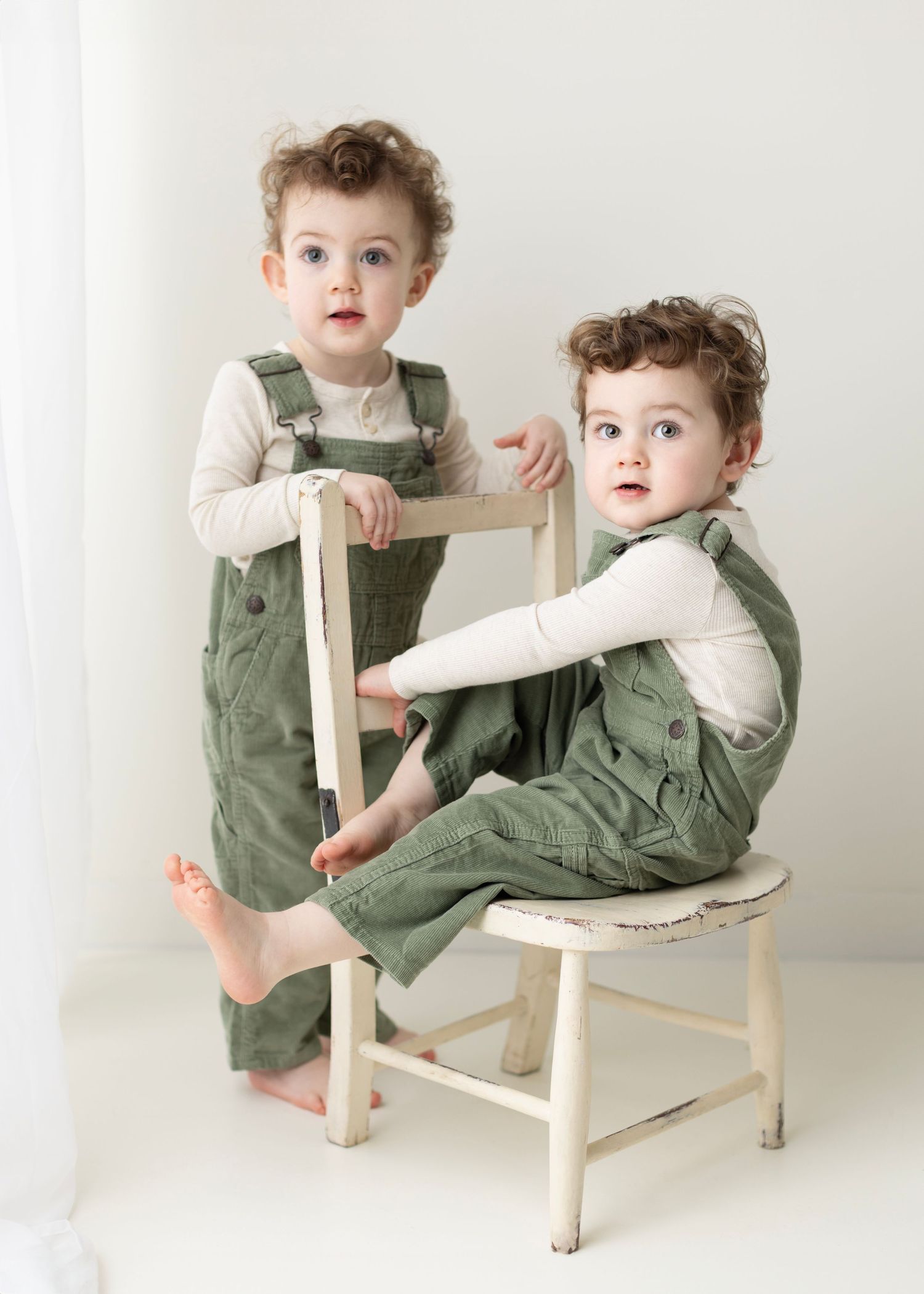 Two children in matching sage green overalls and white shirts pose together on a vintage white wooden chair.