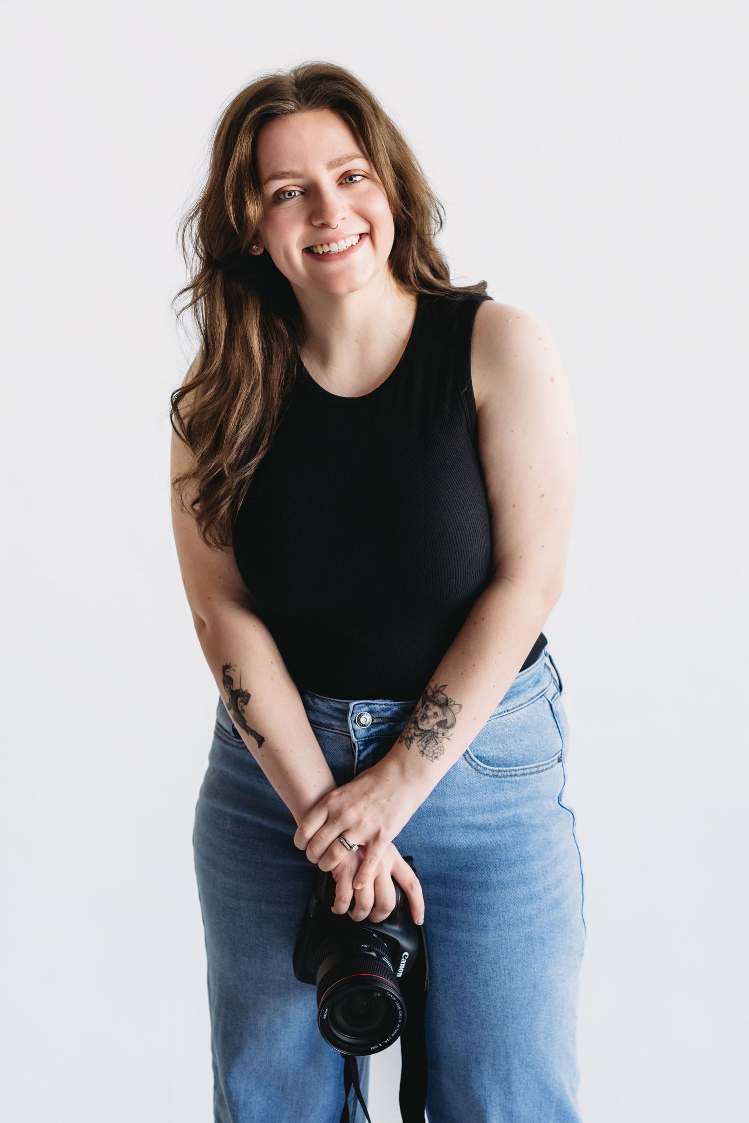 Professional photographer in black sleeveless top and jeans holds camera while posing against white backdrop.