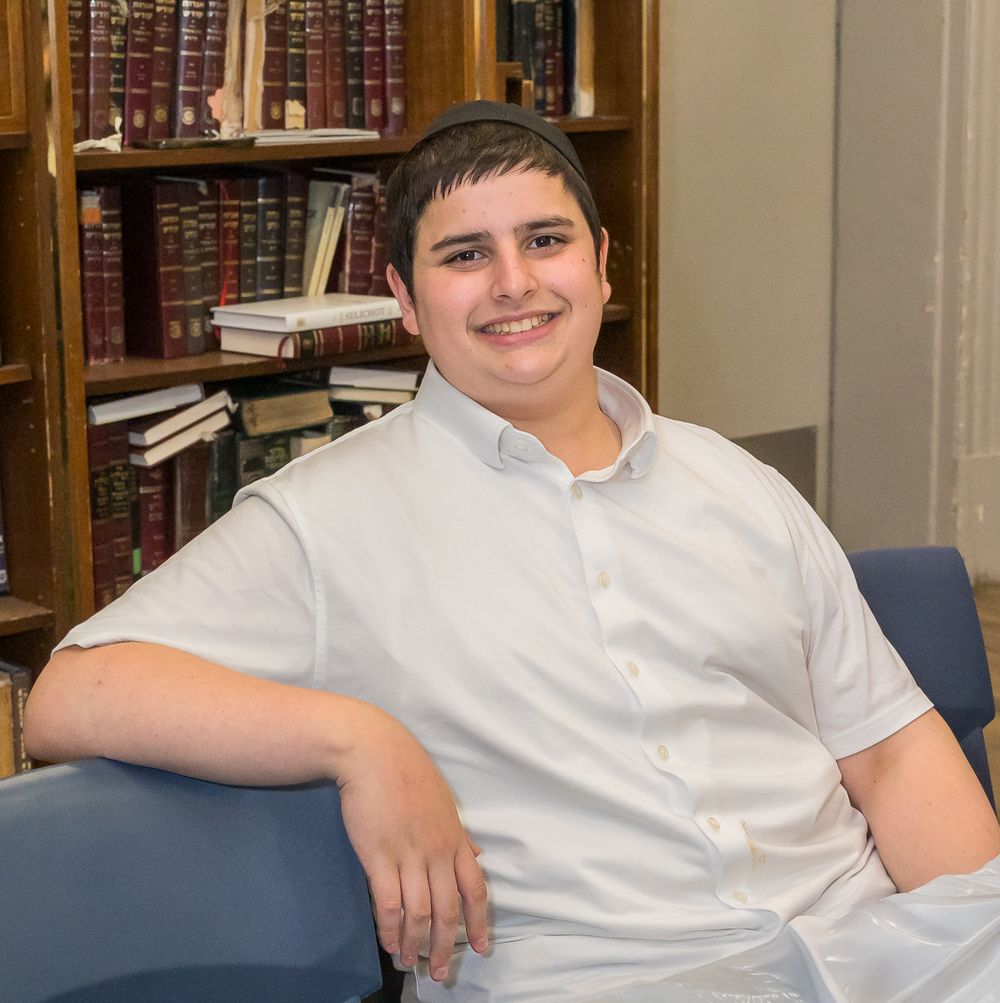 Someone in a white button-up shirt sits casually in a library or study area with bookshelves visible in the background.