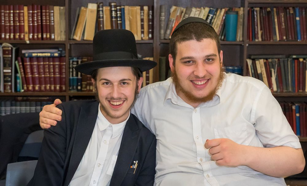 Two men sit together smiling in front of a library bookshelf, one wearing a black hat and suit, the other in white.