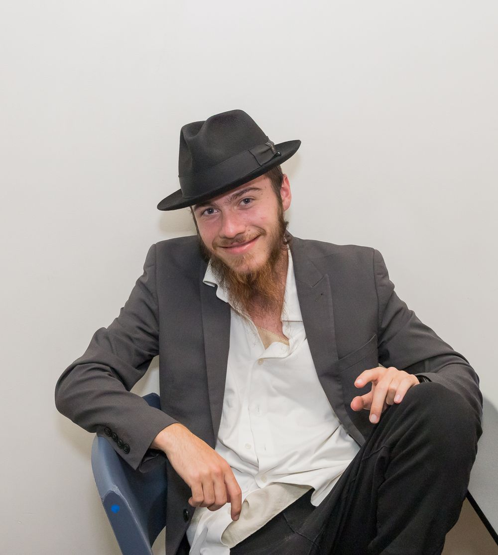 A person in a gray suit and black hat sits casually in a chair while smiling at the camera.