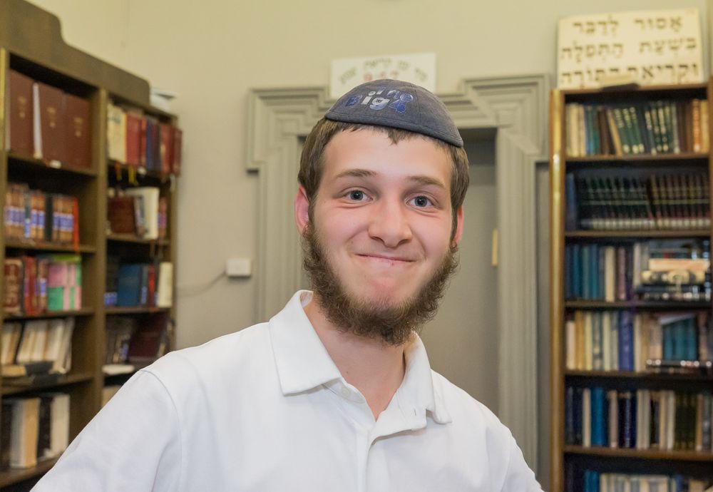 A student in a yarmulke smiles in a library with bookshelves and Hebrew text visible in the background.