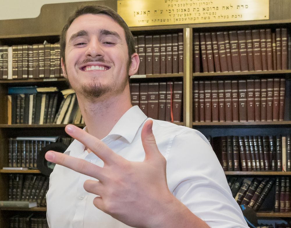 Student gestures with hand while standing in front of library bookshelves filled with bound volumes.