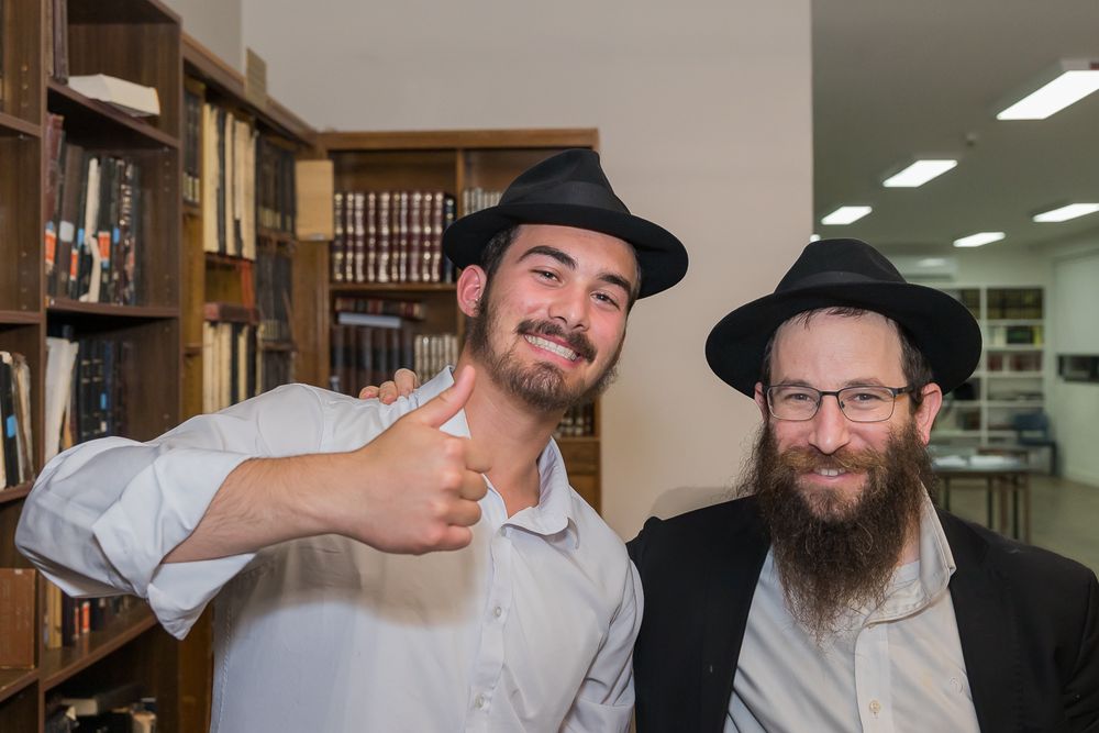 Two men wearing black hats stand together in a library, one giving a thumbs up while the other has a long beard.
