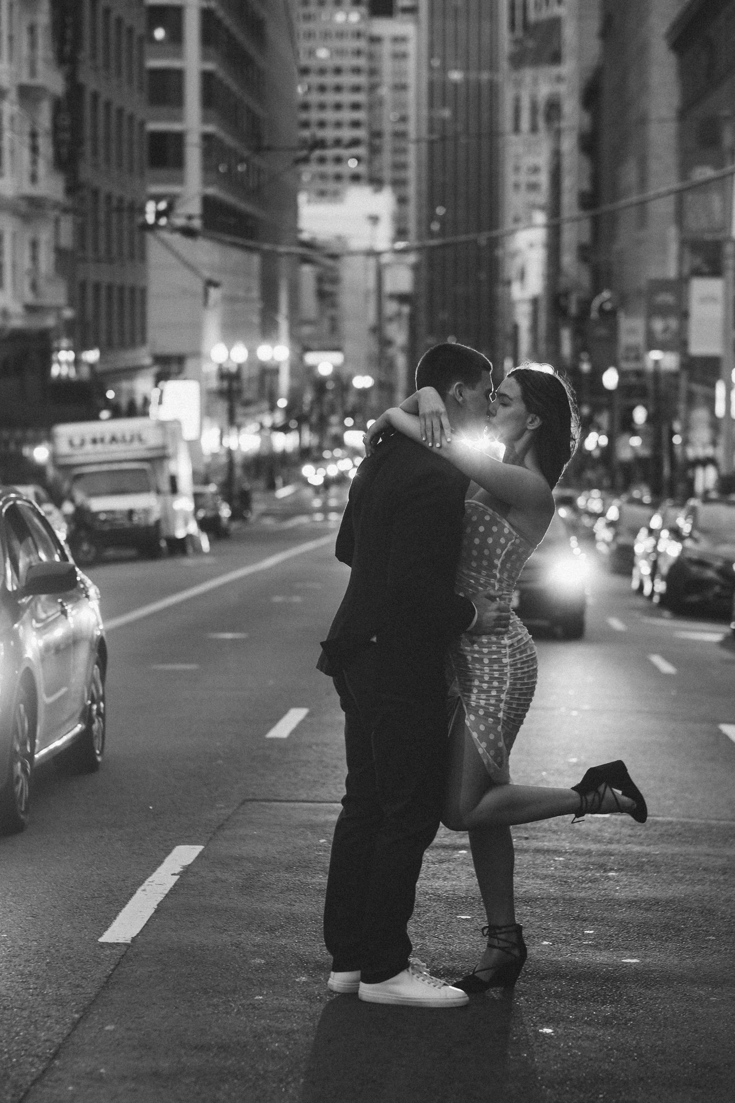 A romantic couple embraces and kisses on a city street at night with urban lights and buildings in the background.