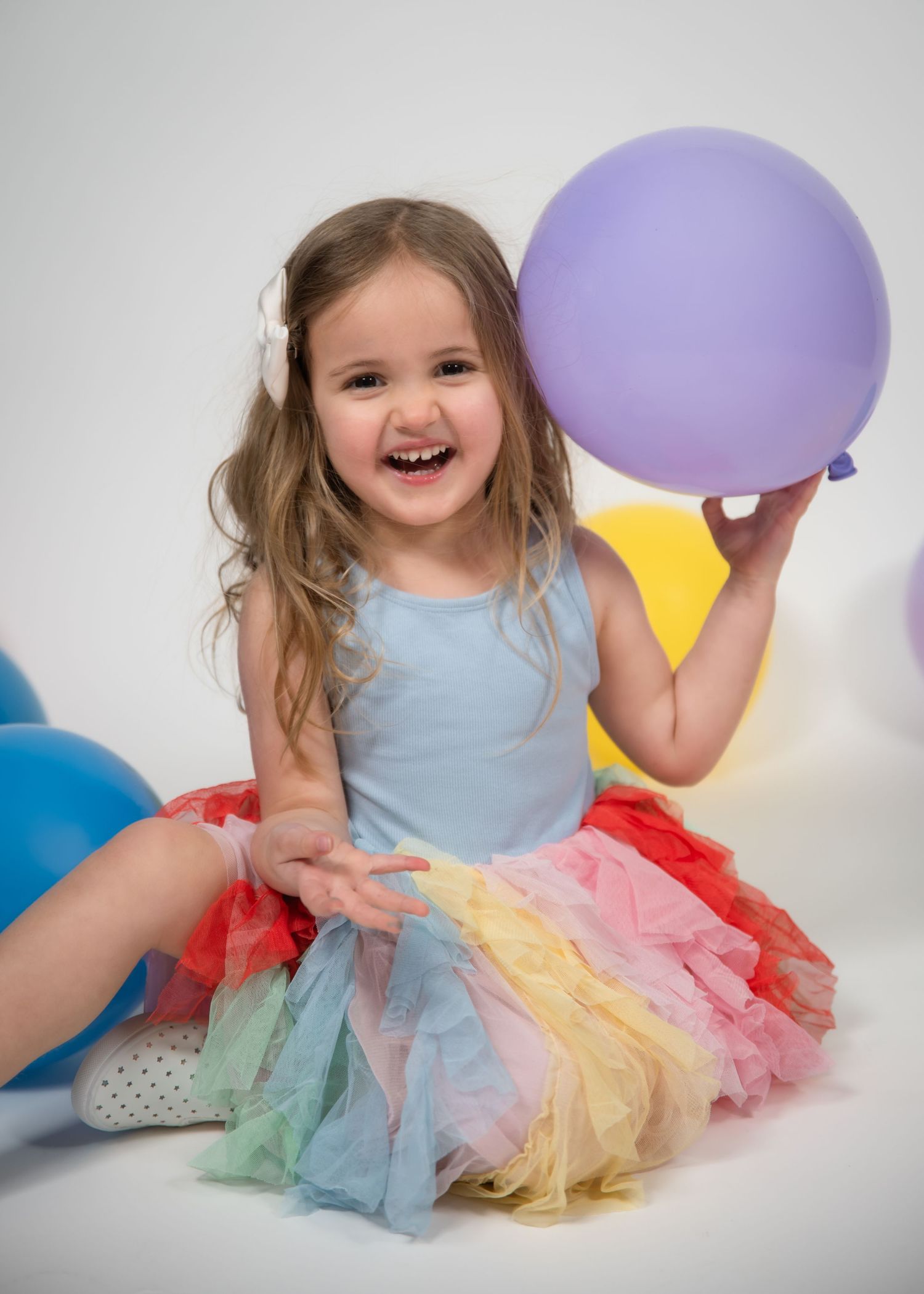 A colorful rainbow tutu dress paired with balloons creates a whimsical studio portrait series against a white backdrop.