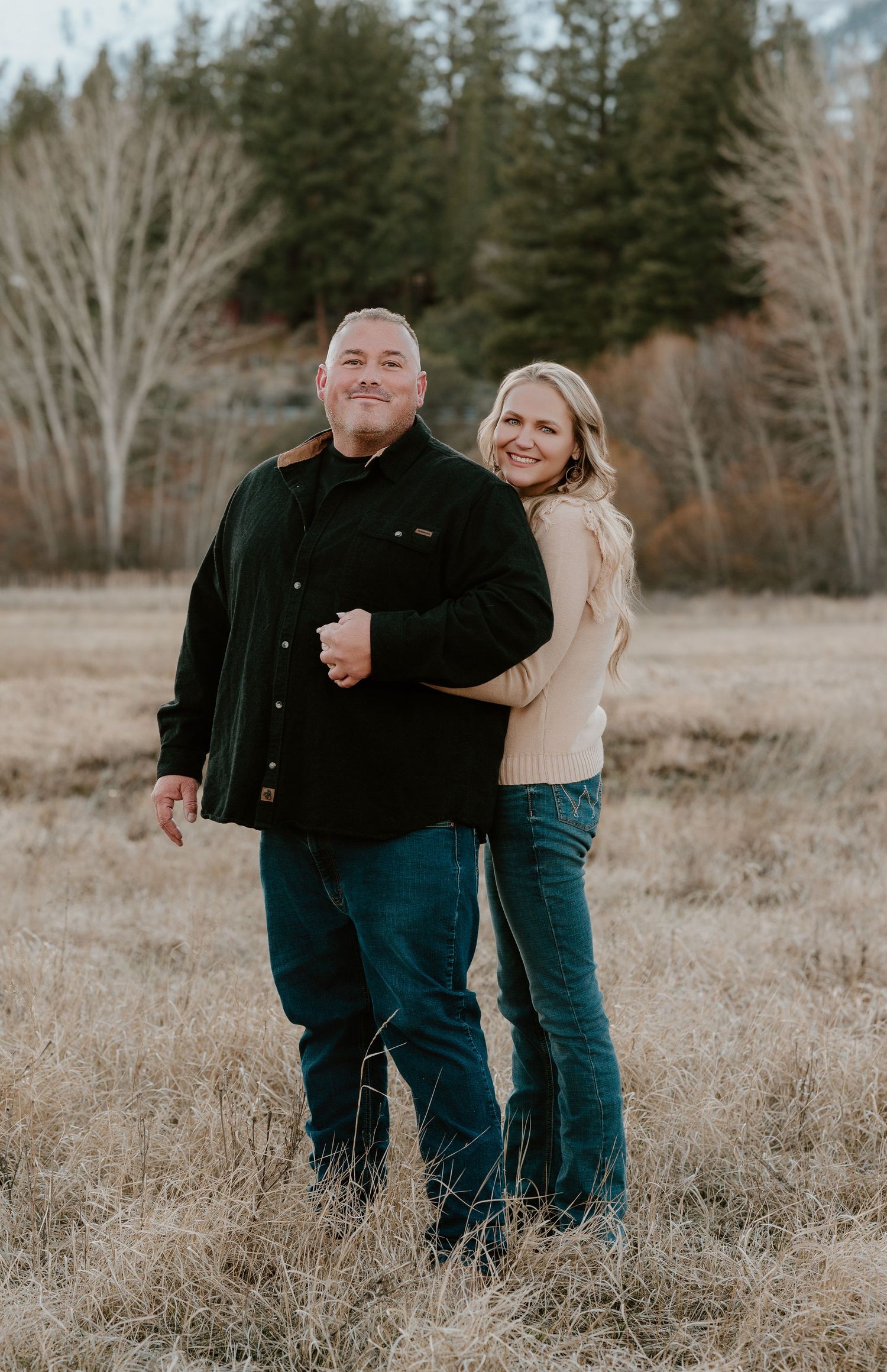 A couple stands together in a field with evergreen trees and bare branches visible in the background on an autumn day.