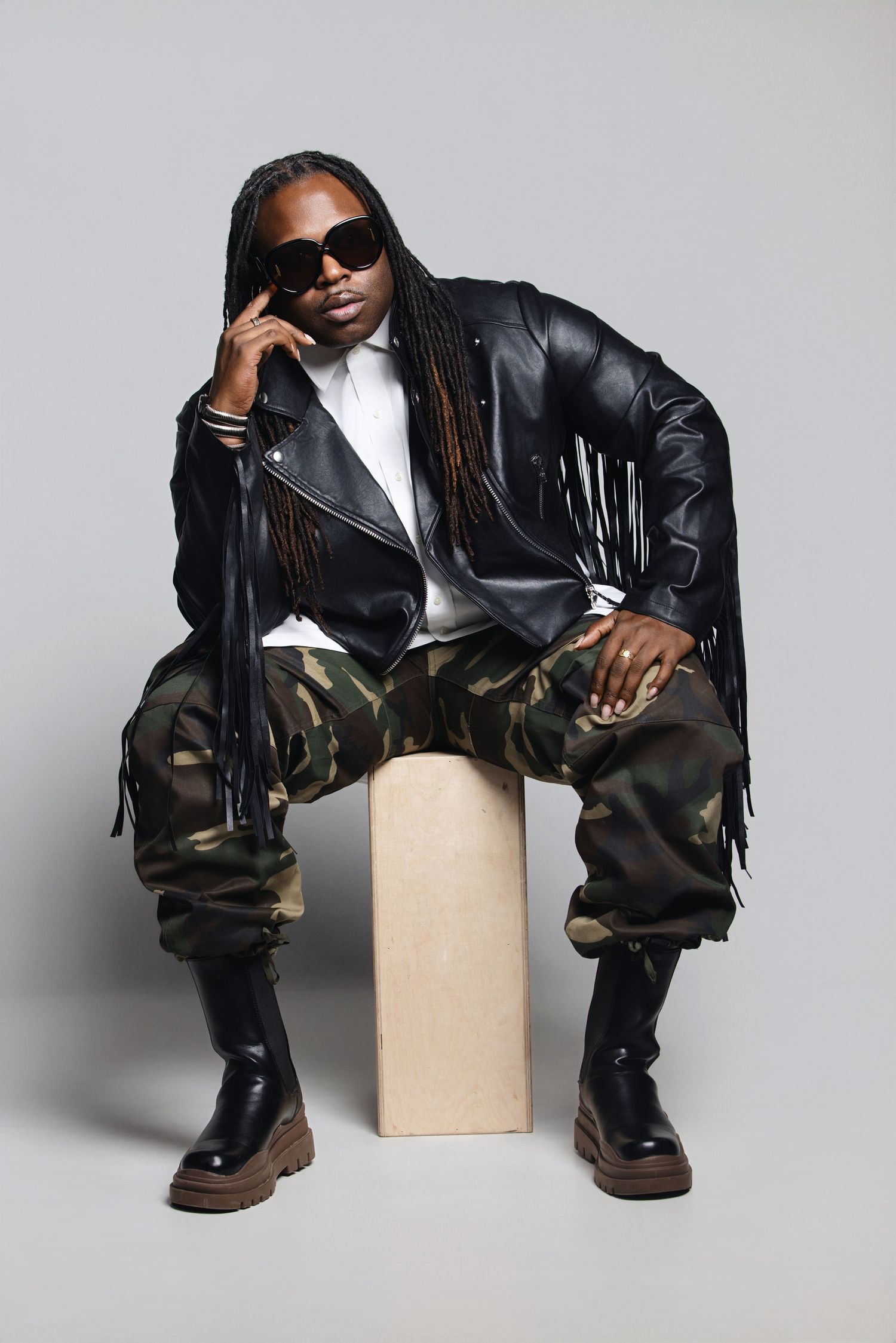 Model poses on cube wearing black leather jacket, camo pants and sunglasses in fashion studio photoshoot.